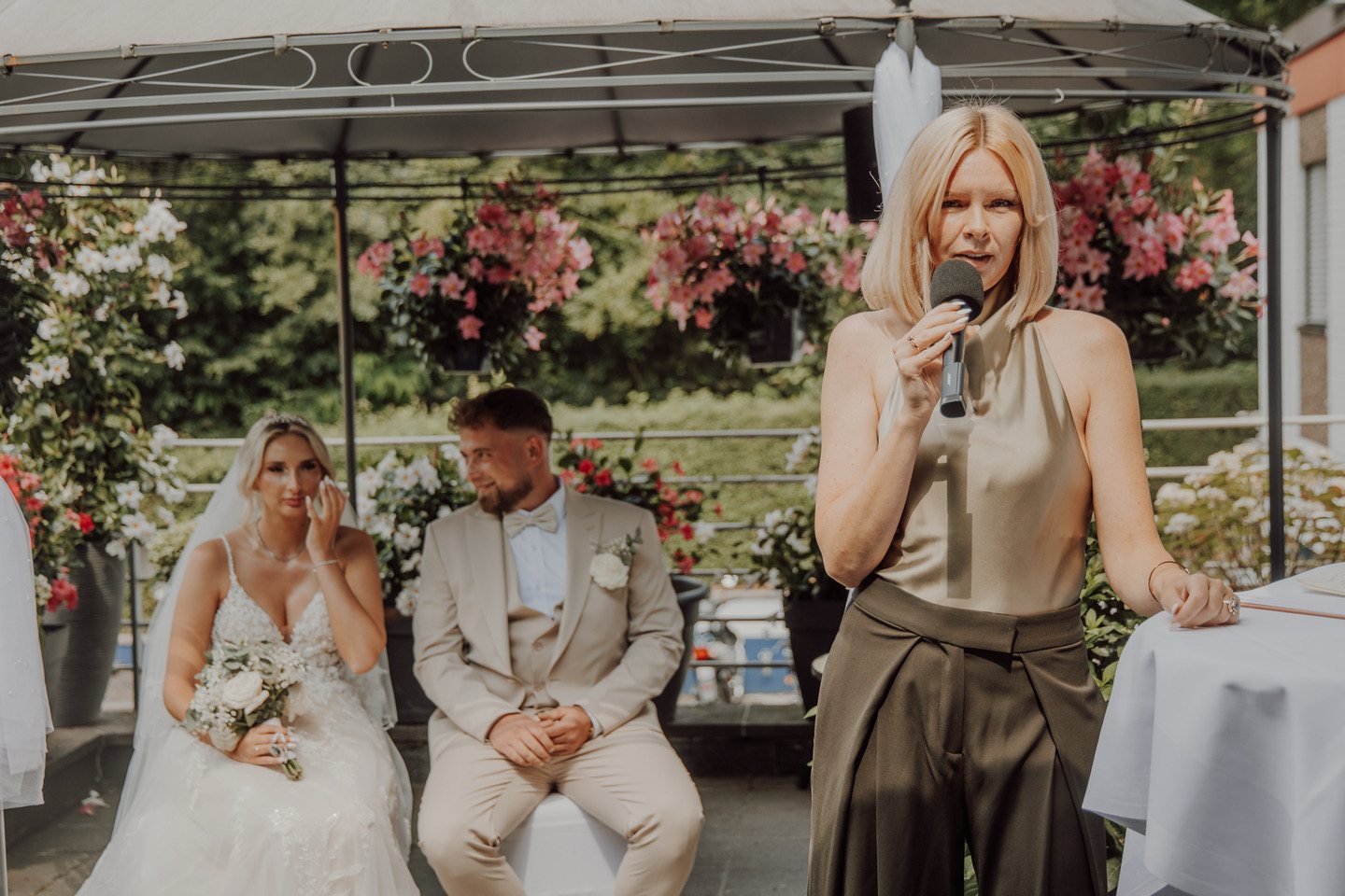 Daria Pfeiffer spricht am Mikrofon, dahinter sitzt das Hochzeitspaar, die Braut wischt sich eine Träne aus dem Gesicht